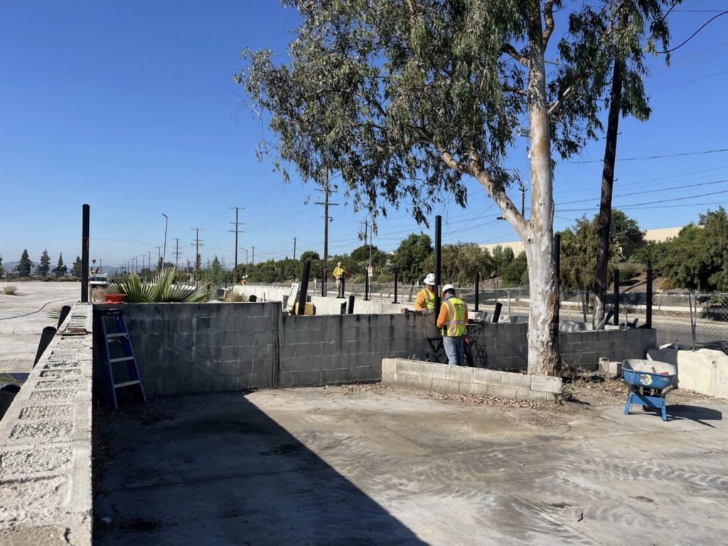 Construction workers in yellow vests and helmets work on a cinder block wall near a tree on a clear day. A blue wheelbarrow and ladder are nearby.