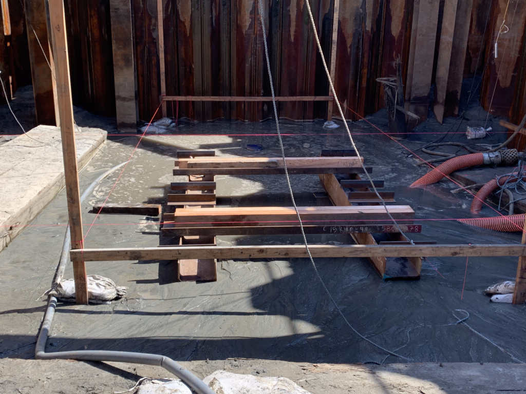 Construction site of Alamitos Bay Pump Station with wooden framework, red guide strings, vertical metal beams, coiled red pipes, and concrete ground