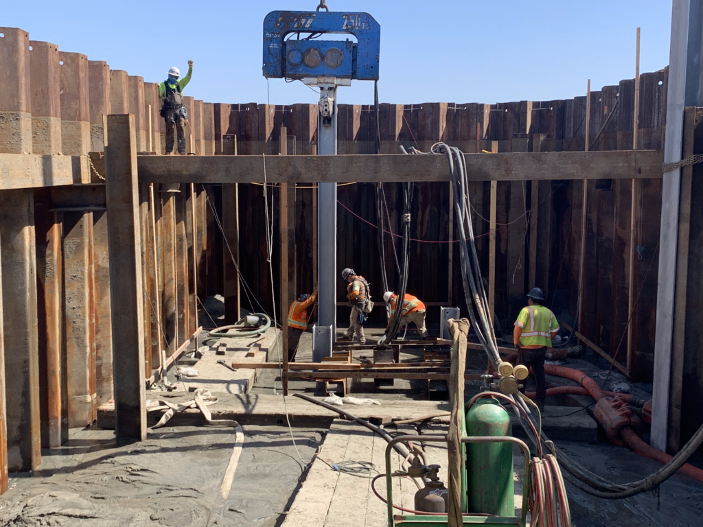 Construction site with workers connecting a steel beam, held by a blue crane, to its base