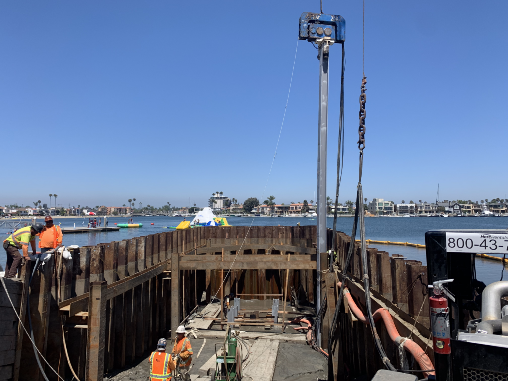 Construction site by the water with workers in safety gear. Crane holding up a steel beam