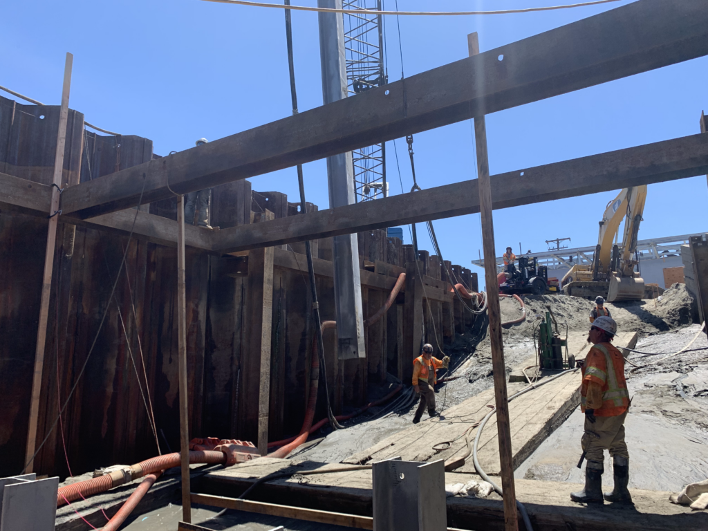 Construction site with workers, steel sheet piles, crane and excavator under a clear blue sky