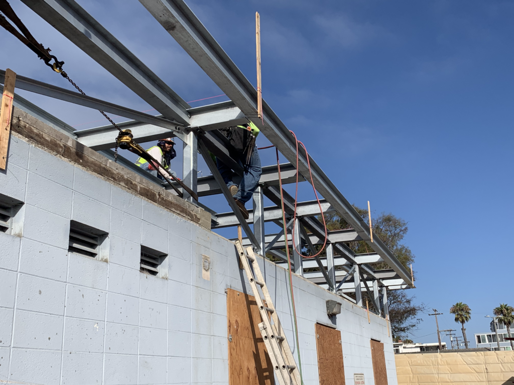 Construction site of a pump station with three workers in safety gear; one climbing a ladder, two working on the roof frame