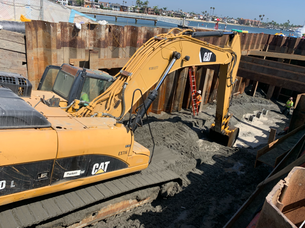 The image shows a construction site at Alamitos Bay with a large yellow CAT backhoe actively working to dig up sediment.