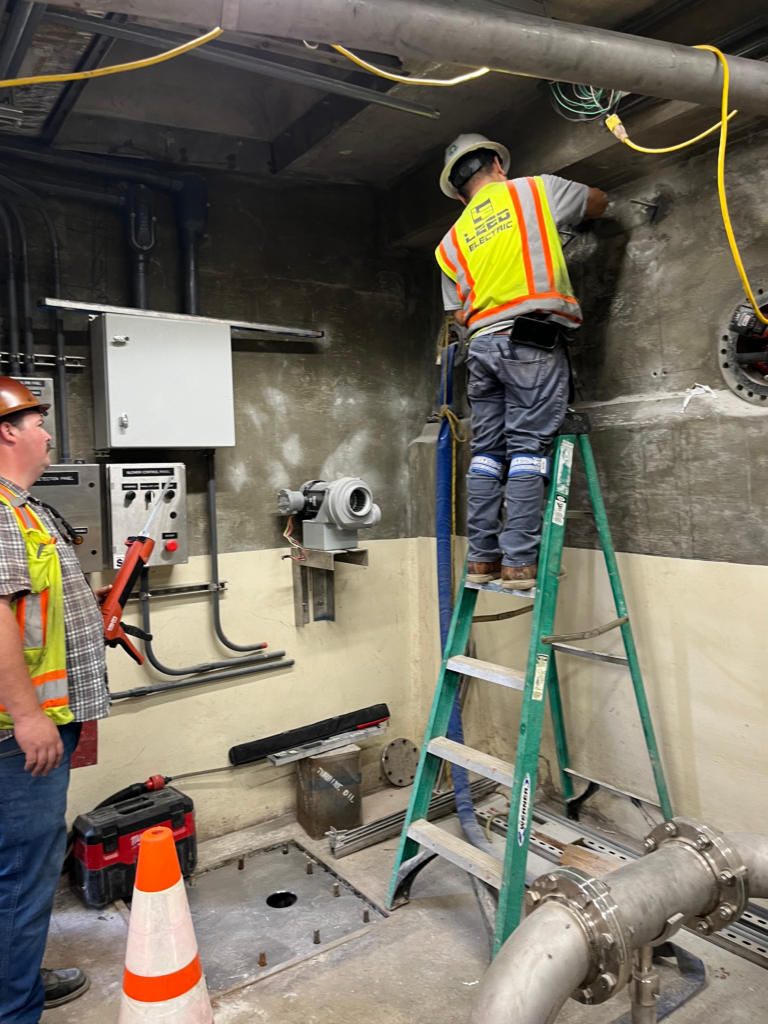 Two men in hardhats are engaged in wall work; one is on a ladder and the other is poised with a chalking gun.