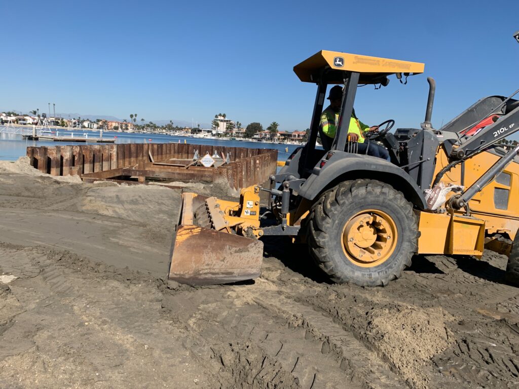 A yellow bulldozer flattens the ground at Alamitos Bay Pump Station, set against a beach backdrop.