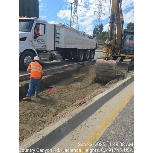 A construction worker in an orange safety vest and hard hat stands in a trench next to a road, with a large white dump truck and a yellow excavator in the background under power lines, indicating ongoing road construction or utility work. The image also includes a timestamp and location: "Sep 23, 2025 11:48:08 AM, Country Canyon Rd, Hacienda Heights, CA 91745, USA."