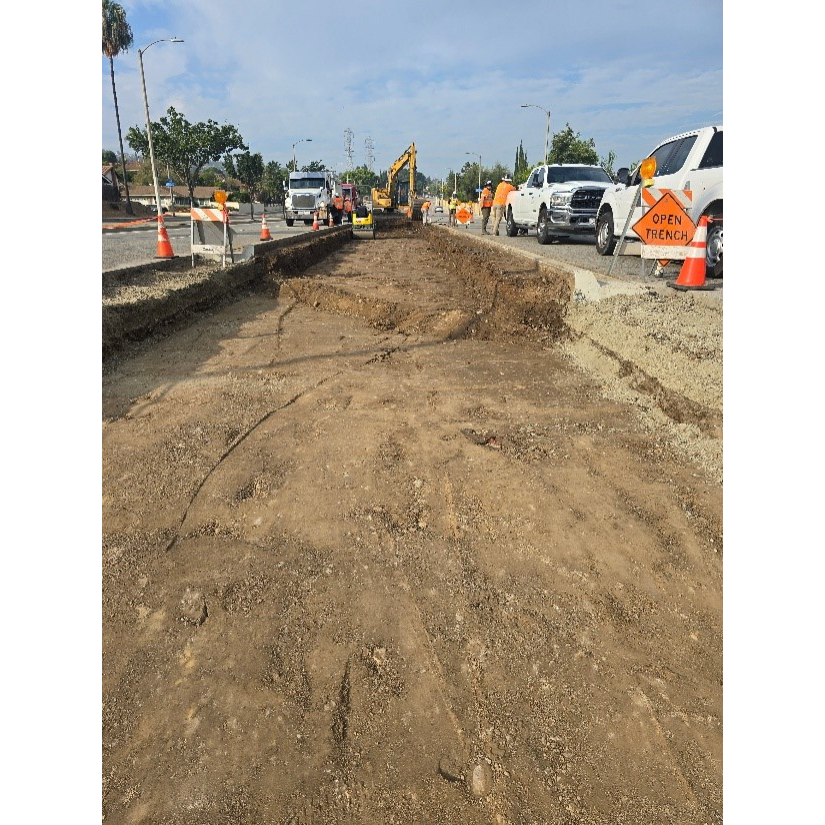 An aerial view of a construction site showing an open trench in the foreground, with construction vehicles like an excavator and trucks in the background, and workers in safety vests. An orange sign with "OPEN TRENCH" is visible on the right.
