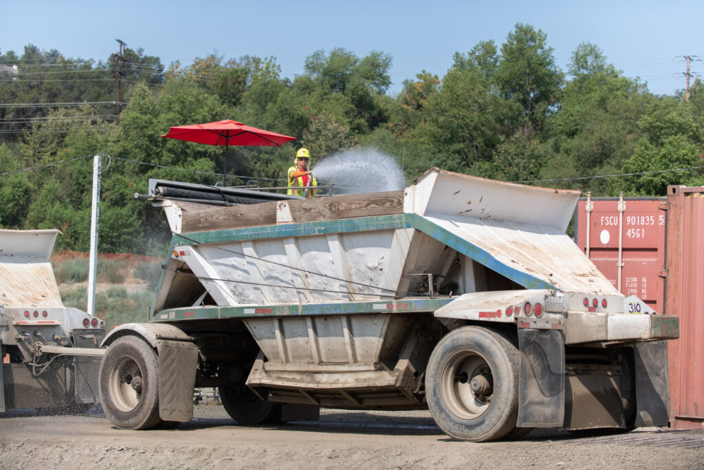 Construction vehicle carrying workload and worker watering down the cargo.